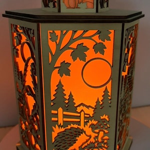 Könnte beinhalten: Eine sechseckige Holzlaterne mit lasergeschnittenen Paneelen, die herbstliche Szenen mit Igeln, Kürbissen, Bäumen und einem Vollmond zeigen. Das Innere ist mit einem warmen orangefarbenen Licht beleuchtet, das die Ausschnitte betont.