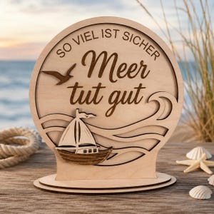 Könnte beinhalten: Ein rundes, dekoratives Holzschild mit einem lasergeschnittenen Segelboot, einer Möwe und stilisierten Wellen. Der Text "SO VIEL IST SICHER" und "Meer tut gut" ist in das helle Holz eingraviert, das auf einem flachen Sockel steht.