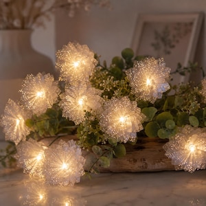 Puede incluir: Una guirnalda de luces decorativas con forma de flor, con pétalos blancos translúcidos y brillantes y centros LED de color amarillo cálido. Las flores están intercaladas con follaje verde y dispuestas en un grupo que emite una luz suave.