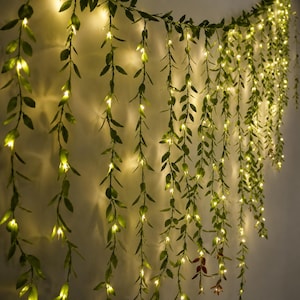 Peut inclure: Un rideau lumineux décoratif composé de multiples brins verticaux de feuilles artificielles vertes entrelacées de petites lumières LED blanc chaud. Les brins pendent vers le bas, créant un effet de cascade de feuillage illuminé sur un mur uni.