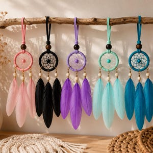 May include: Five small dreamcatchers hanging in a row, each featuring a circular frame wrapped in coloured cord, a central web with a bead, and three dangling feathers. The colours include pink, black, purple, light blue, and dark blue.