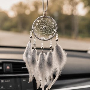 Puede incluir: Un atrapasueños gris colgado de un espejo retrovisor, con un marco circular envuelto en cordón gris, una red tejida con cuentas transparentes, una cuenta plateada central y cinco plumas grises y blancas unidas con cuentas de metal plateado.