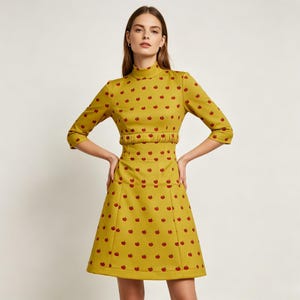 May include: A mustard yellow knee-length dress featuring a high neckline, three-quarter length sleeves, and a gathered waistline. The entire garment is covered in a repeating pattern of small, dark red apple icons arranged in a grid-like formation.