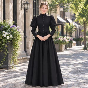 Puede incluir: Un vestido negro largo hasta el suelo con cuello alto, una hilera de pequeños botones en la parte delantera central y mangas largas con hombros abullonados. El corpiño es ajustado y se une a una falda amplia y plisada hasta el suelo.