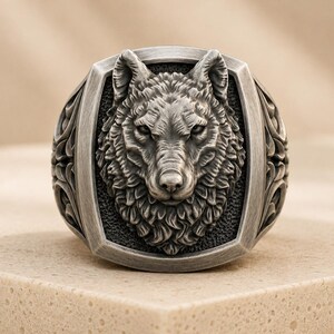 May include: A silver-toned ring featuring a detailed, three-dimensional wolf head relief centered on a rectangular face. The band displays intricate, carved scrollwork patterns along the sides, with a dark, oxidized finish highlighting the textures.