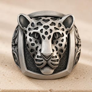 May include: A silver-toned metal ring featuring a sculpted leopard head design. The leopard face is detailed with dark, recessed spots and textured features, set within a rectangular frame that has a brushed metallic finish on the sides.