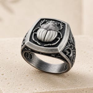 May include: A silver-toned ring featuring a raised, detailed scarab beetle emblem on a rectangular face. The band and sides of the ring display intricate, dark-oxidized scrollwork patterns, providing a high-contrast, textured appearance.