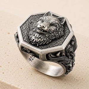 May include: A silver ring featuring a hexagonal face with a detailed relief of a wolf head. The band is engraved with the number 925, indicating sterling silver content. The metal has an oxidized finish to highlight the intricate fur textures.