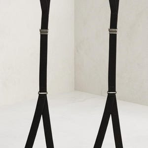 May include: A pair of black suspenders featuring adjustable elastic straps, metallic silver-toned hardware, and four sturdy clips at the base. The design includes a Y-back configuration with a single top loop and two split bottom ends for attachment.