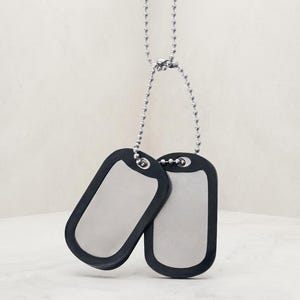 May include: A pair of silver-toned metal dog tags suspended from a silver ball chain. Each rectangular tag features a smooth, reflective center surface framed by a thick, matte black rubber silencer border, creating a high-contrast appearance.
