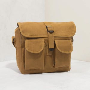 May include: A tan canvas messenger bag featuring a large fold-over flap with a central fabric strap and buckle closure. The front of the bag includes two vertical exterior pockets with rounded flaps, providing additional storage space for items.