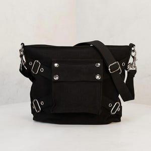 May include: A black canvas shoulder bag featuring a central front pocket with four silver-toned metal rivets. The bag includes adjustable side straps with silver-toned metal buckles and eyelets, along with a wide black shoulder strap attached by metal clips.