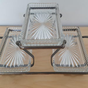May include: A three-tiered serving stand featuring three rectangular clear glass trays with a sunburst pattern on the base. The trays are held by a dark metal frame with two curved handles on the sides, providing a tiered arrangement for display.