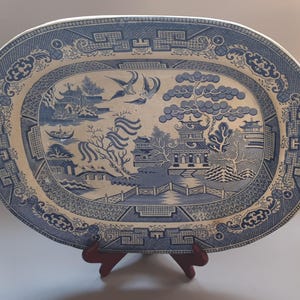 May include: An oval ceramic platter featuring a blue and white willow pattern. The design depicts a landscape with pagodas, a bridge, willow trees, and two birds in flight. The border is decorated with intricate geometric and floral motifs.