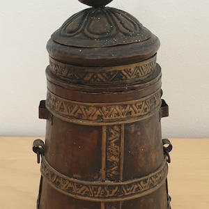 May include: A cylindrical brown metal container with a removable lid featuring a pointed finial. The exterior is decorated with embossed geometric patterns and horizontal bands, and includes small metal loops attached to the sides of the body.