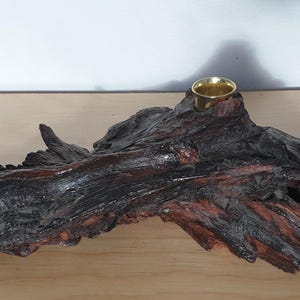 May include: A dark brown, textured piece of driftwood shaped into a candle holder. The wood has a glossy, polished finish with deep reddish-brown undertones. A small, circular, metallic gold-colored cup is embedded into the top of the wood.