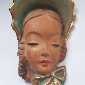 Könnte beinhalten: Eine Wandplakette mit einem skulptierten Gesicht mit geschlossenen Augen und roten Lippen. Die Figur trägt eine große Haube mit türkisfarbenem Innenfutter und Goldrand sowie eine passende türkisfarbene Schleife unter dem Kinn in matter Optik.