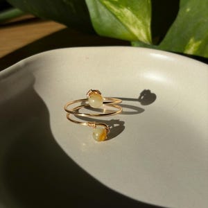 Könnte beinhalten: Ein goldfarbener Drahtring im Bypass-Design mit zwei kleinen, runden, blassgelben Perlen an den Enden. Der Draht ist um die Basis jeder Perle gewickelt und bildet eine filigrane, offene Struktur, die sich um den Finger legt.
