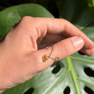 Op de afbeelding: Een goudkleurige draadring met een enkele kleine, ronde, oranje kraal. De draad is gevormd in een open, verstelbaar ontwerp met opgerolde uiteinden, waarvan er één de oranje kraal vasthoudt in een cirkelvormige zetting op de ring.