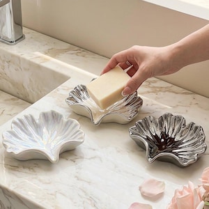 Op de afbeelding: Drie ginkgobladvormige zeepbakjes met een glanzende afwerking. Eén bakje is iriserend wit, één is metallic zilver en één is metallic donkergrijs. Een rechthoekig blokje gebroken witte zeep wordt in het zilverkleurige zeepbakje geplaatst.