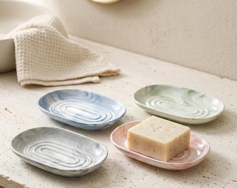 May include: Four oval soap dishes with a marbled finish in blue, green, gray, and pink. Each dish features a concentric oval pattern in the center. A rectangular bar of soap with a bee emblem sits on the pink dish, which has a smooth surface.