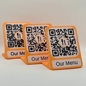 Peut inclure: Trois supports orange en forme de L, chacun doté d'un insert carré blanc avec un QR code noir, une icône centrale représentant une fourchette et un couteau, et le texte Our Menu imprimé en noir en bas. Les supports sont disposés en rangée.