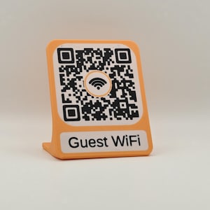 Peut inclure: Un support en forme de L de couleur pêche présentant un code QR noir et blanc avec un symbole Wi-Fi central. Sous le code QR, une étiquette rectangulaire blanche affiche le texte noir "Guest WiFi" dans une police sans empattement.