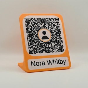 May include: An orange L-shaped stand featuring a white square panel with a black and white QR code. The QR code contains a central circular icon of a person silhouette. Below the QR code, a white rectangular label displays the text Nora Whitby.