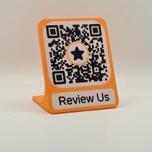 Peut inclure: Un support de présentation orange en forme de L arborant un code QR noir et blanc avec une icône d'étoile noire au centre. Sous le code QR, une étiquette rectangulaire blanche affiche le texte noir Review Us en police sans empattement.