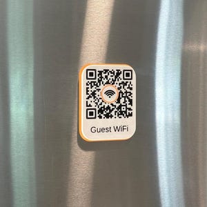 May include: A rectangular sign with rounded corners featuring a black and white QR code, a central orange Wi-Fi symbol, and the text Guest WiFi printed in black. The sign has a white face with an orange border and is mounted on a metallic surface.