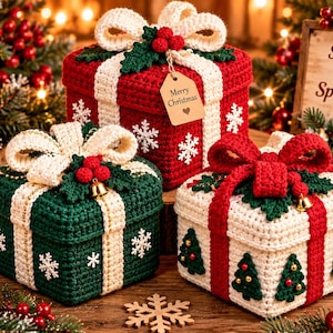 Peut inclure: Trois boîtes cadeaux carrées en crochet aux couleurs rouge, vert et crème, surmontées d'un grand nœud en laine, de feuilles de houx et de baies. Les boîtes présentent des motifs de flocons et de sapins, avec une étiquette Merry Christmas.