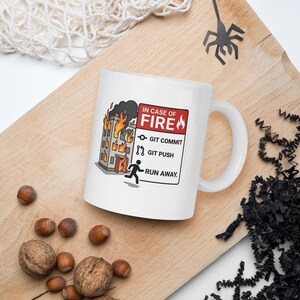 Könnte beinhalten: Ein weißer Keramikbecher mit einer Grafik eines brennenden Gebäudes und dem Text: IN CASE OF FIRE, GIT COMMIT, GIT PUSH, RUN AWAY. Das Design zeigt eine Strichmännchen-Figur, die vor den Flammen flieht, sowie Symbole für Git-Befehle.