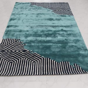 Peut inclure: Un tapis rectangulaire présentant une section centrale aux tons bleu sarcelle et turquoise chinés. Les coins supérieurs et inférieurs sont ornés d'un motif géométrique noir et blanc audacieux composé de lignes angulaires et de losanges.