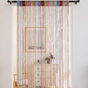May include: A multi-colored string curtain hanging from a black rod, featuring vertical strands in various hues including red, blue, green, yellow, and purple. The thin, individual threads create a semi-transparent, textured partition across the doorway.