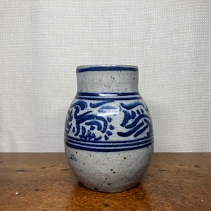 May include: A small, rounded ceramic vase with a speckled light gray finish. The vase features horizontal blue bands at the top and middle, with a hand-painted blue abstract wave or scroll pattern encircling the center section of the body.