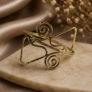 Può includere: Un bracciale a fascia in metallo color oro con una texture martellata. Il design è composto da una struttura in filo continuo che forma due motivi a spirale alle estremità, collegati da linee geometriche angolari che creano una forma aperta.