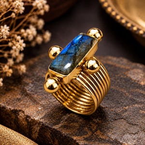 May include: A gold-toned ring featuring a rectangular labradorite gemstone with blue iridescence. The setting includes four polished gold spheres at the corners of the stone, and the band is constructed from multiple thin, parallel gold-toned wires.