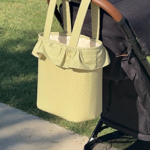 May include: A light green tote bag with a white polka dot pattern, featuring a ruffled trim around the top opening and two fabric handles. The bag is hanging from the handle of a black stroller, showing a cream-colored interior lining.