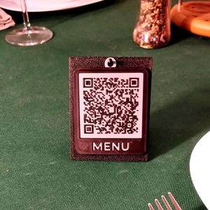 Può includere: Un supporto rettangolare nero con un inserto quadrato bianco contenente un codice QR nero e la parola MENU stampata in lettere maiuscole bianche nella parte inferiore. Il supporto presenta una superficie testurizzata e un piccolo foro.