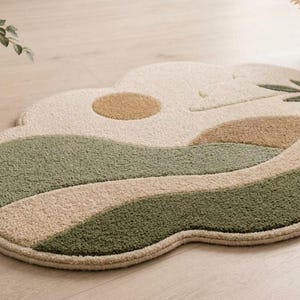 May include: An irregularly shaped area rug featuring a tufted texture with a landscape design. The rug displays a color palette of cream, sage green, and tan, depicting a sun, rolling hills, and a leafy branch motif across its surface.