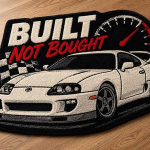 Könnte beinhalten: Ein getufteter Teppich mit einem weißen Toyota Supra Sportwagen, einer schwarz-weißen Zielflagge und einem roten Drehzahlmesser. Der Text BUILT NOT BOUGHT ist in fetten weißen und roten Buchstaben auf schwarzem Grund dargestellt.