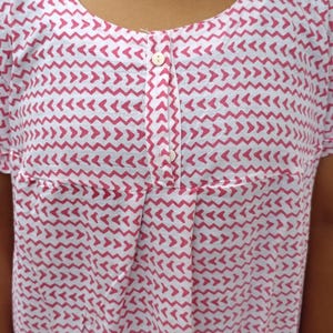 May include: A white short-sleeved top featuring a repeating pink chevron pattern. The garment has a round neckline and a central placket with two white buttons. Below the placket, there are vertical pleats that add texture to the fabric.
