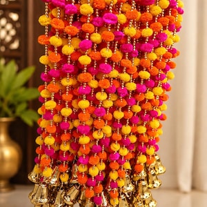 Puede incluir: Una guirnalda decorativa colgante con múltiples hilos verticales de pompones de colores amarillo vibrante, naranja y rosa intenso, intercalados con pequeñas cuentas doradas. Cada hilo termina en su base con una campana de metal dorado.