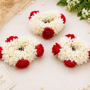 May include: Three circular floral garlands crafted from small white jasmine-like blossoms and accented with vibrant red roses. Each ring-shaped arrangement features a dense, textured pattern of white petals punctuated by several deep red rose blooms.