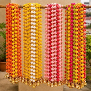 May include: Four vertical garlands hanging from a wooden rod, each featuring rows of small, round, colourful pom-poms separated by gold-toned beads, with each strand ending in a small, metallic gold-coloured bell at the bottom.