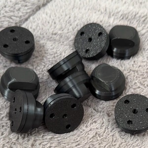 May include: A collection of small, black, circular plastic components. Each piece features a textured top surface with three evenly spaced holes arranged in a triangular pattern, and a smooth, rounded base with a protruding centre section.