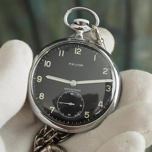 May include: A silver-tone pocket watch with a round black dial, white Arabic numerals, and silver hands. The dial features the text FAVOR and SHOCKPROOF. A small sub-dial for seconds is positioned at the bottom. A metal chain is attached to the top.