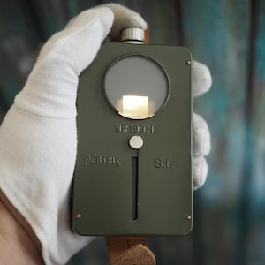 May include: An olive green rectangular metal device with a circular glass lens, a silver top knob, and a vertical sliding switch. The front is embossed with the text PERTRIX, NR 199 M, and S.F. A brown leather strap is attached to the bottom of the unit.