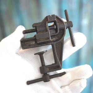 May include: A small, dark gray metal bench vise featuring a C-clamp base with a threaded screw mechanism and a top-mounted sliding jaw. The tool includes two T-shaped handles for tightening the clamp and the jaw, showing a weathered, industrial finish.