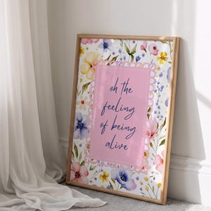 Könnte beinhalten: Ein rechteckiger gerahmter Druck mit einem Aquarell-Blumenmuster in Gelb, Rosa und Blau. In der Mitte befindet sich ein rosafarbenes, gewelltes Rechteck mit dem dunkelblauen kursiven Text: oh the feeling of being alive. Der Rahmen ist aus hellem Holz.